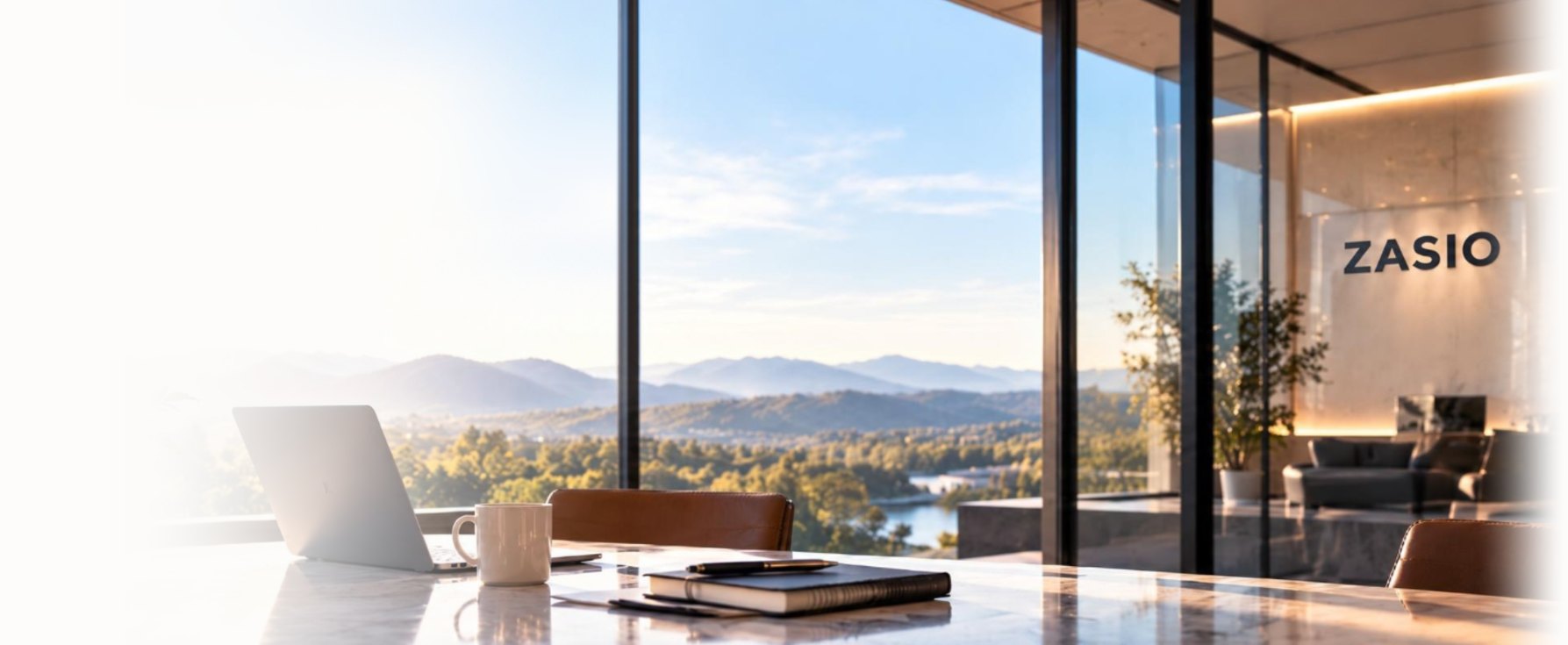 A Zasio office at golden hour: a laptop, coffee mug and notebook on a marble table beside floor-to-ceiling windows looking out over wooded hills and a river, with the Zasio name on the lobby wall
